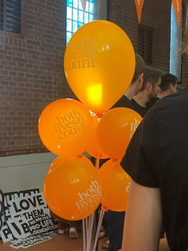 A bunch of orange balloons with 'both lives count' printed on them, as well as a small circular logo featuring a foetus. A pile of signs in the corner includes wording of 'Love them both' (extrapolating) and 'we are the pro-life generation', 'mother & child' and more