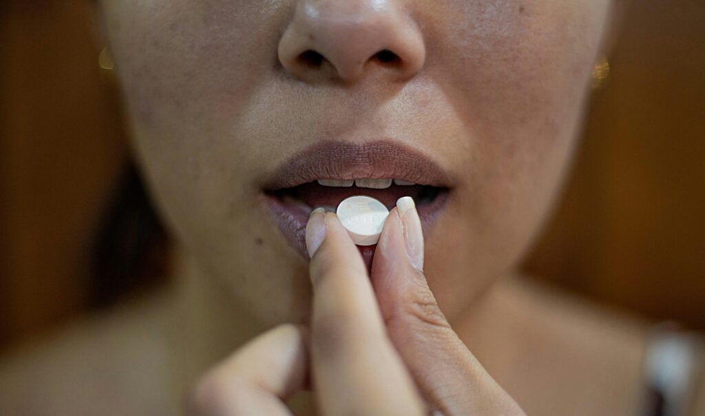 A woman holds a round, white tablet between her thumb and index finger and is putting it into her mouth. We cannot see her eyes, which are out of frame