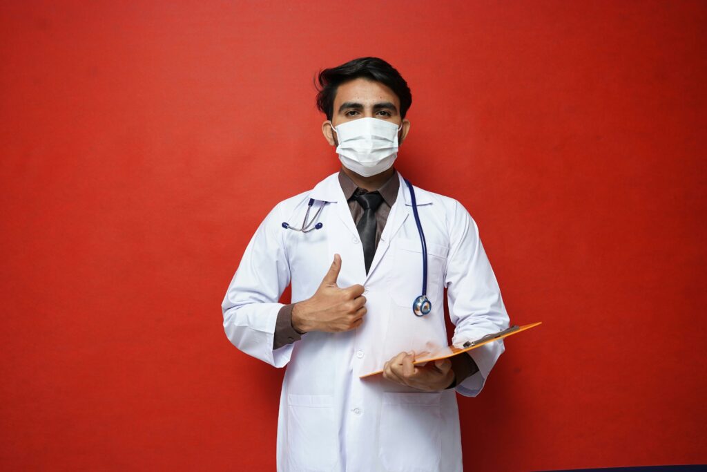 A man with dark, shiny hair wearing a white labcoat and stethoscope around his neck stands in front of a red wall. His right hand is doing a thumbs up and he's holding an orange clipboard in his left. He's wearing a white surgical face mask.