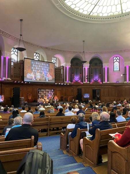 People sitting in the pews of a church repurposed as a conference room, with a panel of four people on stage and a video feed of them projected on a large screen above them