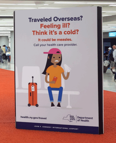 A Department of Health measles alert sign in New York, reading "Traveled Overseas? Feeling ill? Think it's a cold? It could be measles. Call your health care provider" and featuring a brown-skinned cartoon figure holding a tissue and their phone while sitting on an airport bench next to their suitcase, looking worried.