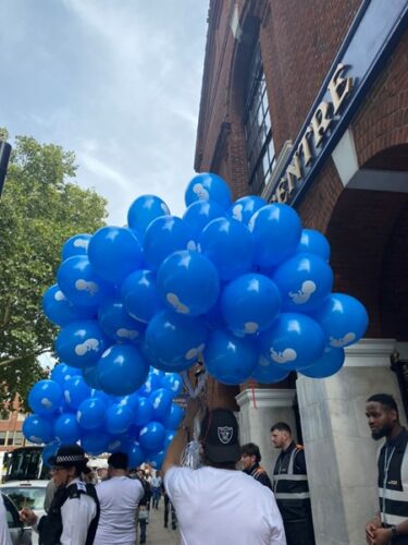 Men on the street in white t-shirts and black baseball caps hold large bunches of blue balloons, each bearing a white printed image of a foetus