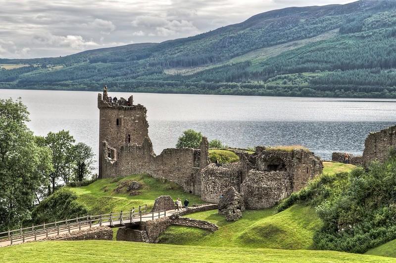 A ruined castle in front of a lake, a stone bridge leading up to it, and green rolling, rising hills in the background. Loch Ness, Scotland.