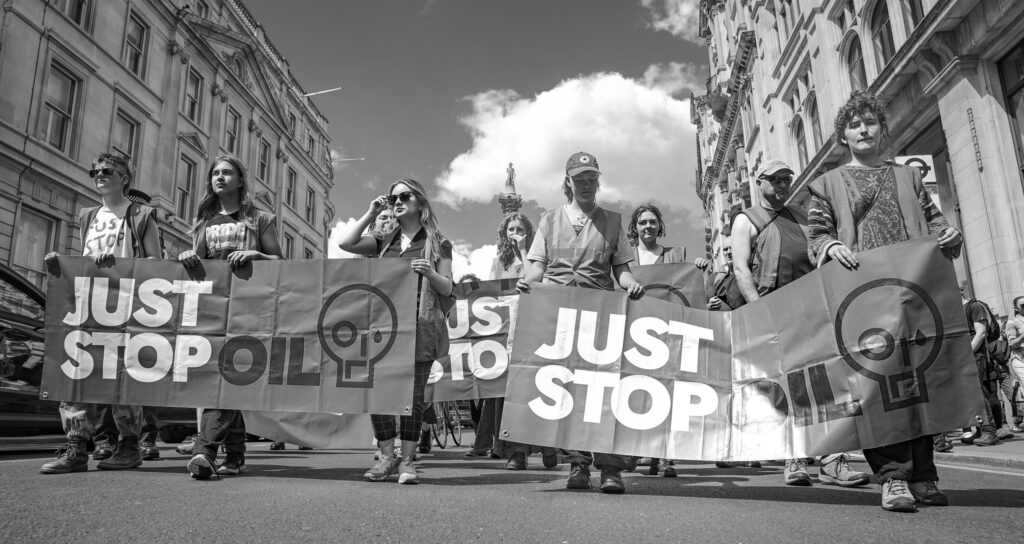 A black-and-white photo of a Just Stop Oil protest on Whitehall, London, UK. 9, mostly female, protesters are walking with three large Just Stop Oil banners.