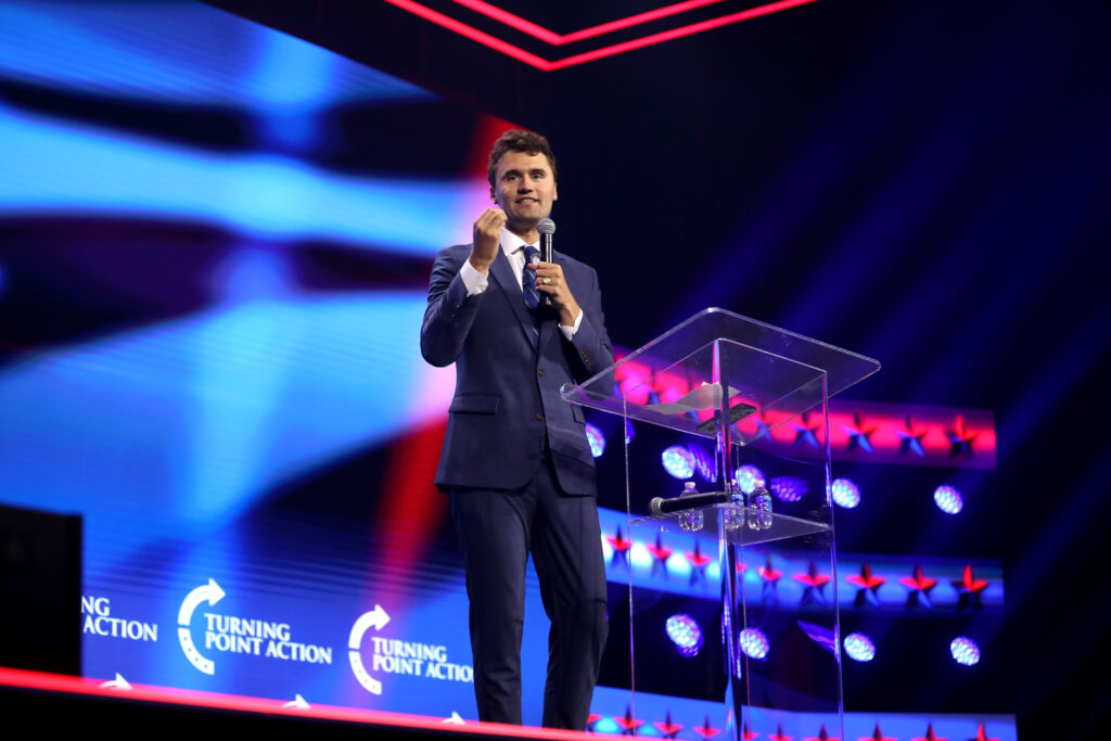 Charlie Kirk onstage with a red and blue-lit stage backdrop that features 'Turning Point Action' logos. He's holding a microphone and standing behind a perspex lectern
