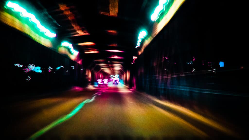 Blurred tunnel and vehicle lights at night, in a range of neon colours, as if the viewer is travelling fast in a car, or perhaps high (or both)