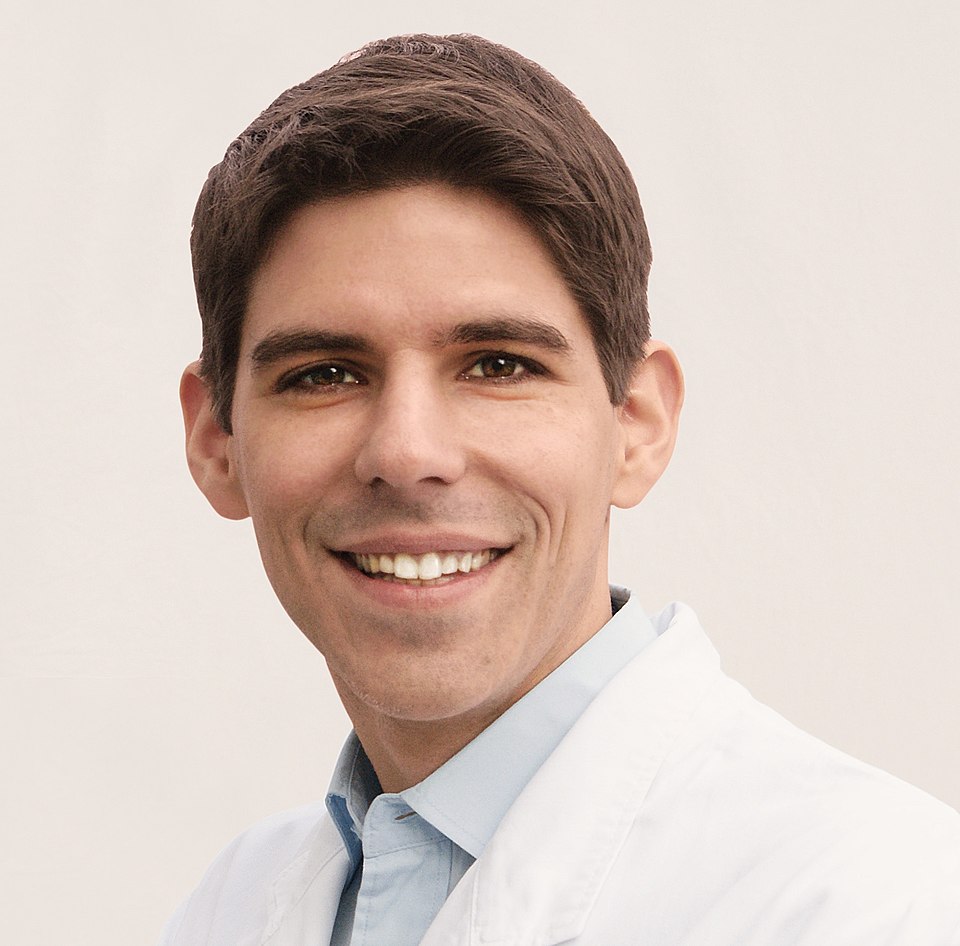 David Bardens, a German physician weather a light blue shirt and white coat, smiles at the camera. He has short, thick brown hair and brown eyes and is clean shaven.