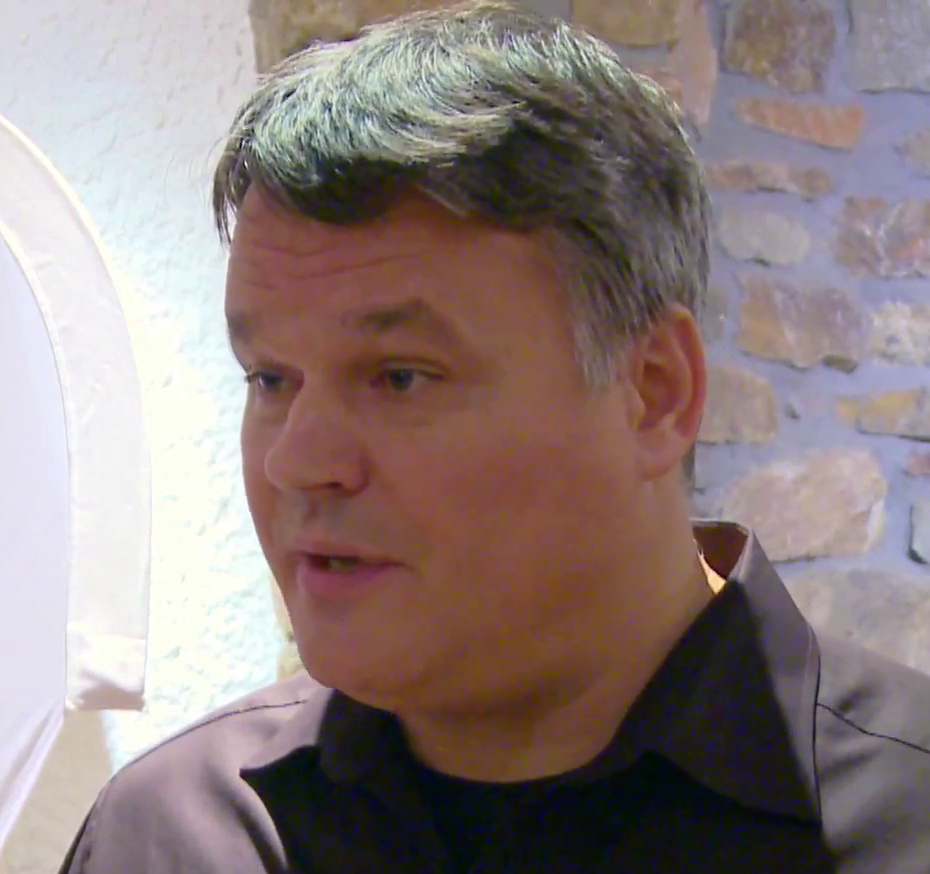 Stefan Lanka speaks to someone in front of him. He's wearing a black shirt over a dark t-shirt and has short, greying hair