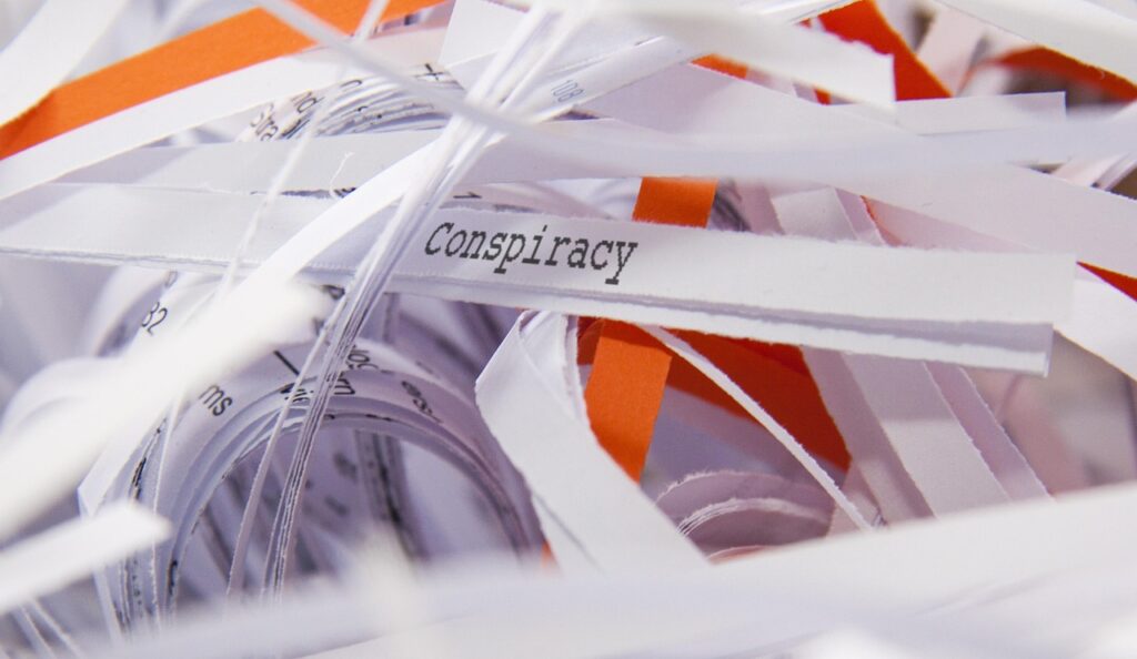a pile of white and orange strips of shredded paper. One strip has the word 'conspiracy' in a typewriter font