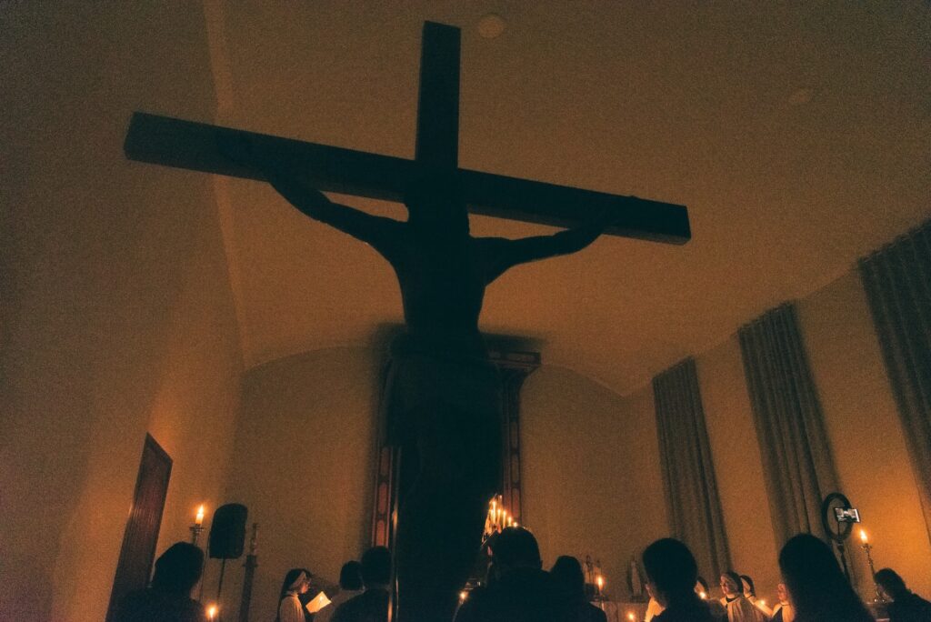 A silhouette of a crucifix in a dark, candle-lit room with some people below it