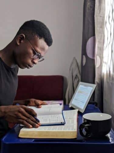 A black man wearing glasses and writing in a notebook with an open book and tablet screen suggesting he is conducting research. 

Photo by Oladimeji Ajegbile on Unsplash