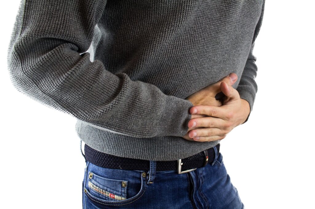 A view of the torso of a male-presenting person who's leaning slightly forward with both their hands clasped over their navel, appearing to be in discomfort or pain. They're wearing a grey jumper and blue jeans with a black belt.