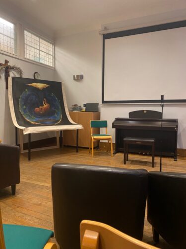 A small lecture room containing an upright piano and stool below a pull-down projector screen, a microphone stand, some chairs and a large piece of paper or fabric draped above a table, on which is printed a foetus in a blue, round space that looks like an oubliette.
(Credit: Abi Kennedy)