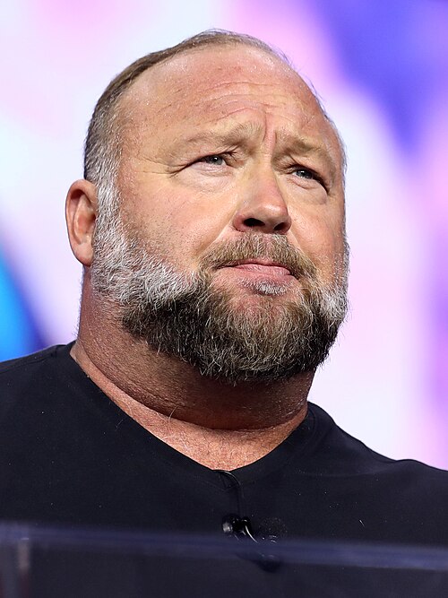 A close-up of Alex Jones standing at a lectern in front of a bright display, wearing a black t-shirt and looking serious, with a grey and dark-brown beard and moustache. 
