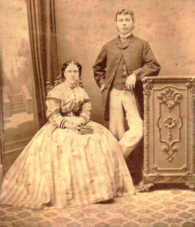 A sepia photograph depicting a woman seated and a man standing beside her, both formally dressed and looking toward the camera. Behind them a backdrop has been painted to give the impression of a doorway opening out into a garden.
The woman is wearing a long-sleeved floor-length striped dress and is holding a book in her lap.
The man is wearing pale trousers, and a darker coat open at the front to show a waistcoat with watch chain.