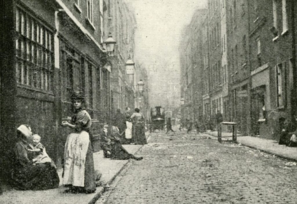 A grainy black and white photograph of a street scene. Brick buildings four storeys tall line both sides of a cobbled street. Horse-drawn vehicles and pedestrians are visible down the street in the background. In the middle distance on the pavement on the left hand side some children are sitting. In the foreground a standing woman looks toward the camera, while a woman sits on the pavement beside her with a child on her lap.