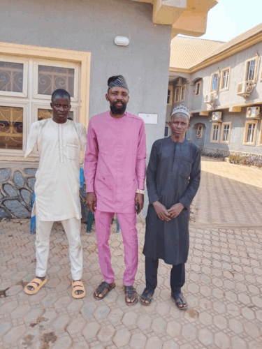 Three black men wearing buba-style white, pink and grey (respectively) Nigerian tunics and sandals. The man in white on the left has no arms as they've been amputated close to each of his shoulders.
