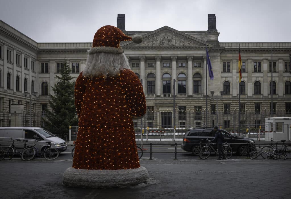 In a Finnish plaza or square there stands a red and white Santa statue/figure, with fluffy robe and crimped white hair, covered in small yellow fairy lights. It faces away from the camera towards a large official-looking building that has some flags outside. An undecorated christmas tree stands to the left. Bicycles are strewn along a railing between Santa and the building, next to the road.