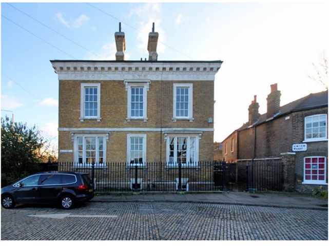 A photograph of a two storey house which is remarkably similar to the illustration, including the wrought iron fence, the appearance of the flat roof, the number of windows, and the number of chimneys. The two chimneys are set close to the centre of the house. There is a tree to the left of the house, and other brick buildings to the right.