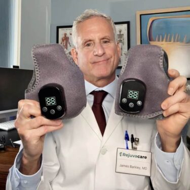In an office, a white-haired man in a white labcoat, off-white shirt and dark red tie holds up two grey felted devices with simple black-plastic, two-number/three-button displays on them. His name badge reads 'Rejuvacare - James Barkley, MD'