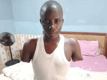 A young black man wearing a white vest sits on a bed, both his arms have been amputated not far below his shoulders.