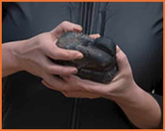 A closeup of the woman's hands from the image above. The object she is holding is amorphous, lumpy and rounded, with some kind of groove in it. Part of it seems smooth and black, elsewhere it appears rough and grey.
The left hand is anatomically unusual with very long fingers. What might be an extra thumb with no clear connection to either hand is partially visible poking up from behind the object.
