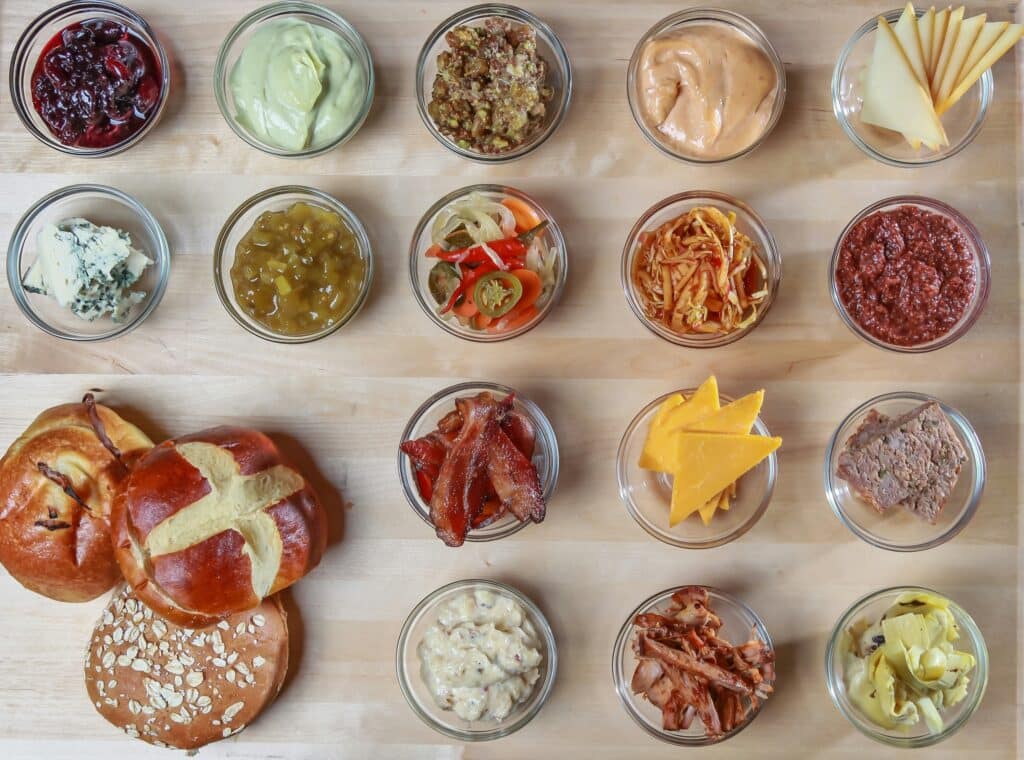 Four rows of a total of 16 small glass bowls of different foodstuffs, including sauces, cheese, bacon and paté, with a sliced round brioche bun, an onion roll and a brown oaty roll piled in the lower left of the image