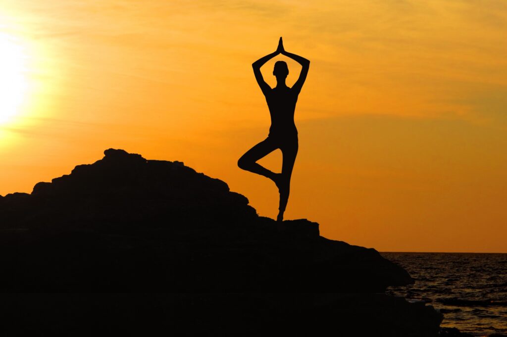 A silhouetted feminine figure on rocky ground strikes a yoga pose, their right foot resting on their left knee and their palms clasped above their head. The sun is rising or setting behind them and there's a body of water off to the right