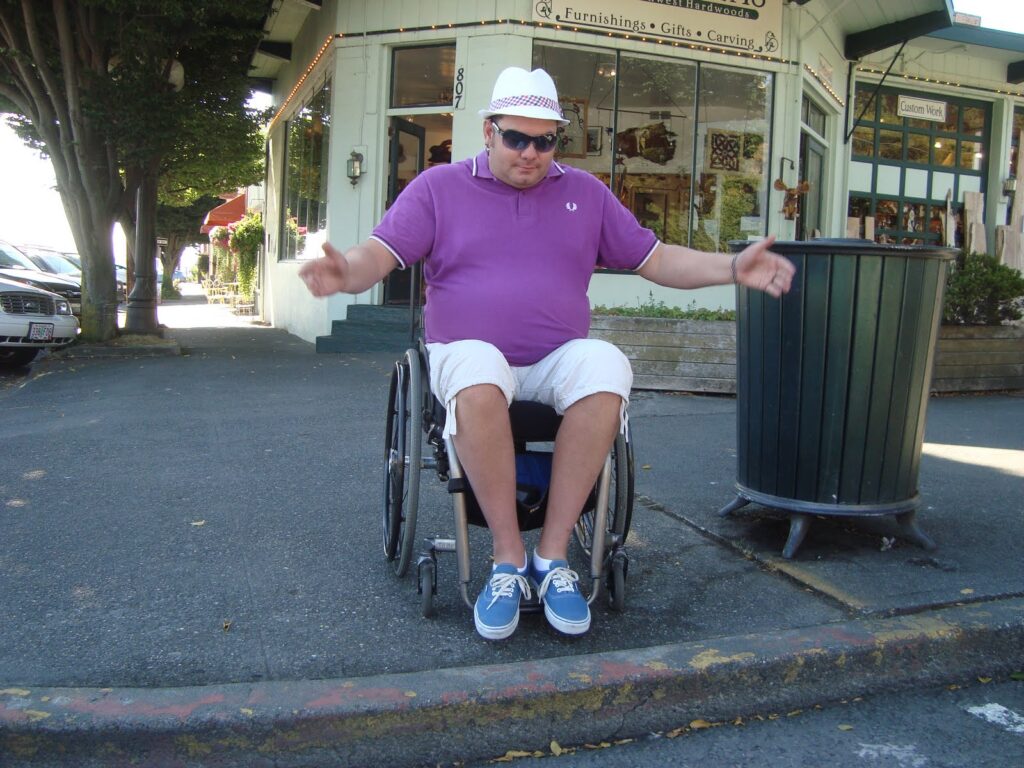 A street corner with shops fronting onto a pavement, then a road. A man seated in a wheelchair is unable to descend from the pavement to cross the road, because the kerb steps down without a ramp. He holds out his hands to the side, as if indicating the problem and a sense of frustration.
