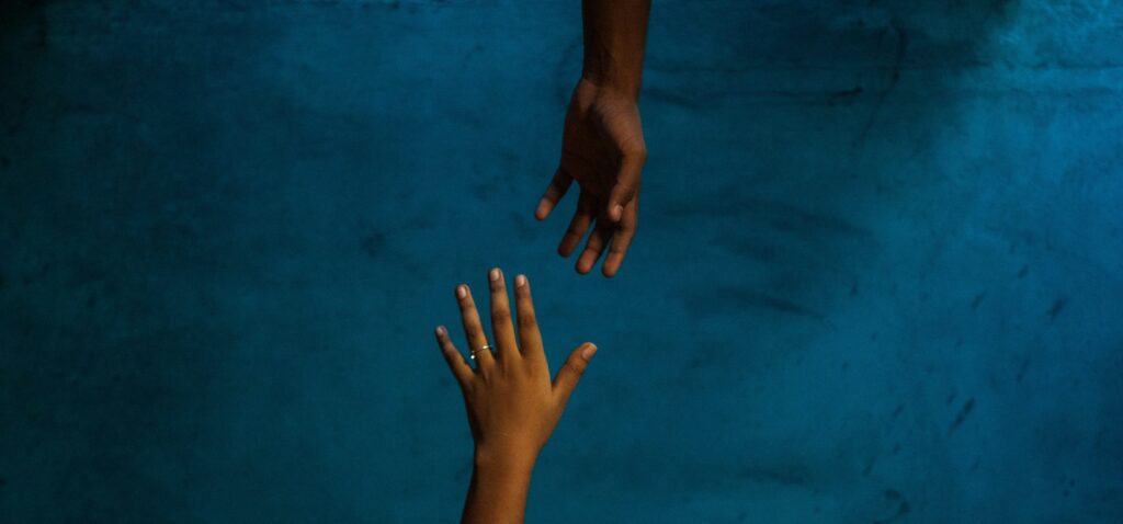 Against a textured, dark blue background, a dark-skinned hand reaches down to meet another, similar hand, reaching upwards.