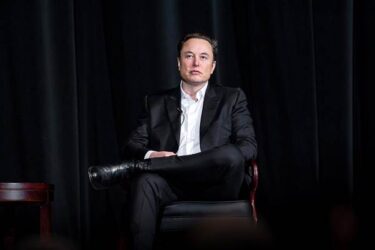 Elon Musk seated, as if on a stage. He has one leg crossed over the other and wears black shoes, black trousers, and a black jacket which is open to show a white shirt. The background is a black curtain.