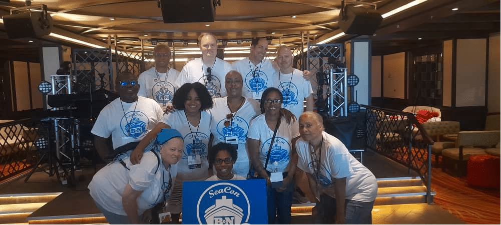 Eleven people stand behind a SeaCon logo board, wearing lanyards, t-shirts and in some cases resting their arms on others' shoulders.