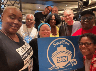 Nine people huddle around a blue and white SeaCon Black Non-Belief project logo placard, featuring a stylised front-on ship and waves inside a circle