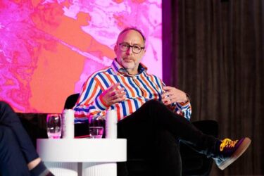 Jimmy Wales seated on stage at a conference. He wears dark shoes with yellow laces, black trousers, and a shirt with broad white, orange and blue stripes. He seems to be talking to someone just out of shot. The background is a bright but indistinct graphic.