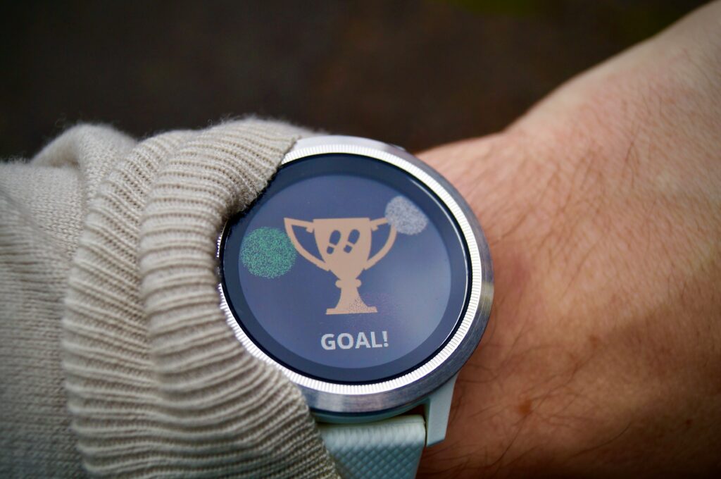 A white man's wrist with a smart whatch visible beside a pushed-back jumper sleeve. The display shows a gold trophy with two footstep icons, two exploding fireworks in the background and the word 'GOAL!' beneath