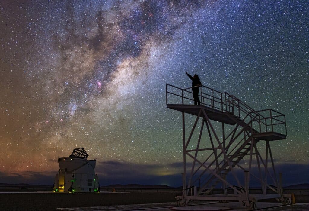 Photograph showing a flat landscape with clear night sky above. Running across the left hand side of the sky is the glowing band of the Milky Way. The rest of the sky contains many individual stars.
In the distance on the ground is a building from which a large astronomical telescope is pointing upwards. In the foreground is a staircase to a raised platform. A person stands on the platform, reaching toward the sky as if to touch the Milky Way.