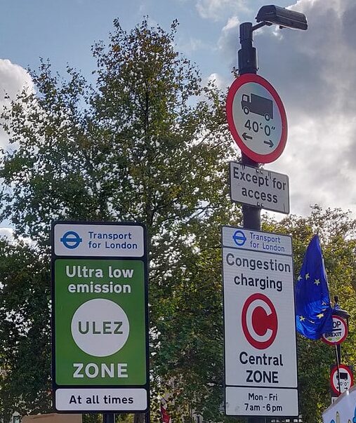 A collection of London road signs, two from Transport for London warning of the Ultra-low emission zone (ULEZ). one for the Congestio charging zone. Some others note large vehicle restrictions. There's an EU flag flying to the right
