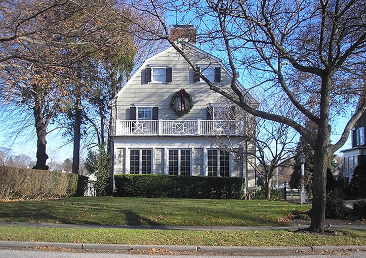 A white and grey-fronted house with a first floor balcony and large wreath hanging in its centre, three ground-floor windows and two on the top two floors, stands behind a long green lawn and short hedge, with leafless trees dotted around 