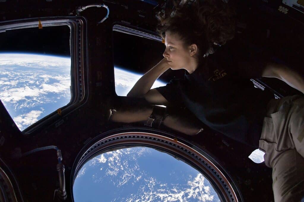 A photograph taken in the dark interior of the International Space Station cupola, where large windows show a view of the sunlit Earth below. Visible in light reflected by Earth, a woman looks out at the view.
