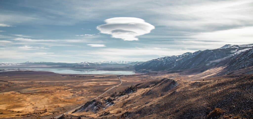 A panoramic view of Mono basin in California, featuring snow-capped mountains and Mono Lake, with a road winding past it, and prominent lenticular clouds floating above in the centre of the image