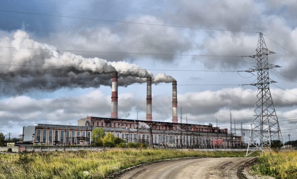 A factory or power plant with three narrow chimneys, all spewing large amounts of grey pollution clouds into the sky. A curving dirt road is in the foreground, leading past an electricity pylon to the right.
