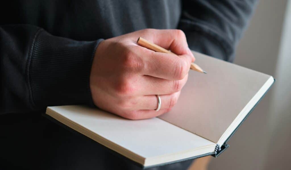 A masculine hand holds a small brown wood pencil ready to mark a small notebook, whose open pages are blank