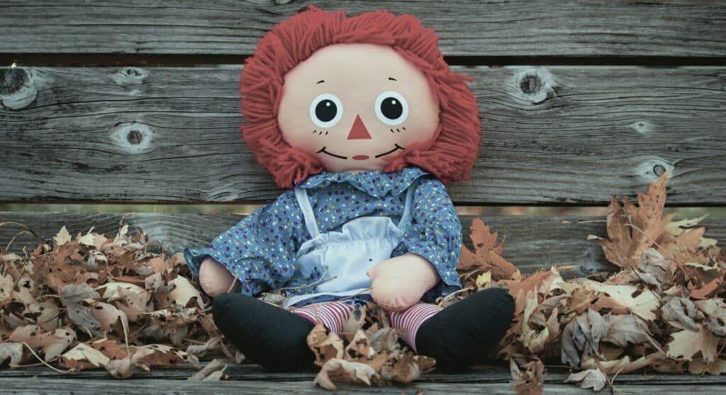 Raggedy Ann - a sewn fabric pale girl doll with bright red stringy hair, a triangular nose and spotted shirt, apron and stripy tights with black shoes - sat on a wooden bench surrounded by fallen autumn leaves