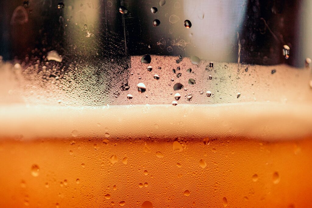 A close-up photograph of a glass of beer, showing the foamy "head" and some of the golden-coloured beer beneath. There are droplets of moisture on the outside of the glass.