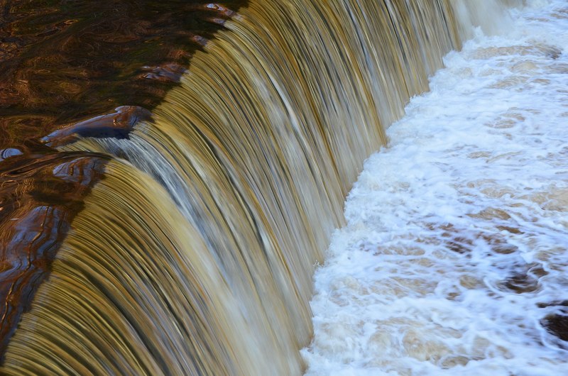 A photograph of water flowing over a weir. Upstream the water appears smooth and placid, while downstream the surface is choppy and covered with swirls of bubbles.