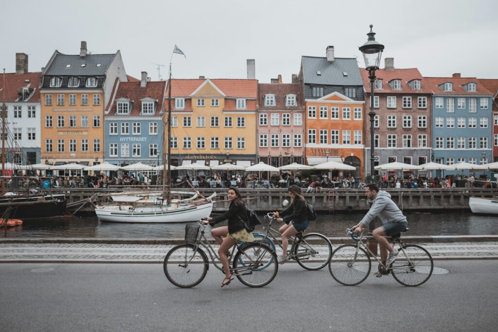 A photograph showing two women and a man riding bicycles along a riverside path. Some small sailing vessels are moored on the far side of the narrow river. On the far river bank there are many awnings with people sitting and standing under them such as at cafes or market stalls. Beyond the awnings is a row of terraced riverfront buildings with steeply pitched roofs, each building painted a different colour.