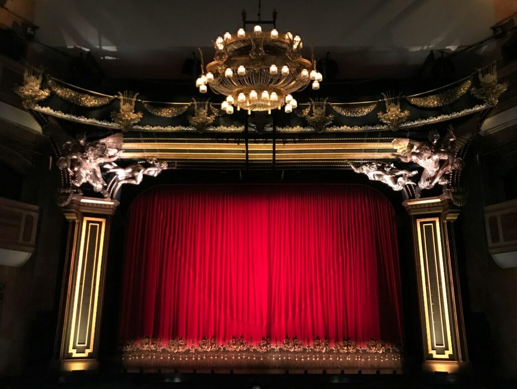A photograph taken inside a theatre, looking toward the stage. The red stage curtain is closed. The proscenium arch framing the stage is richly decorated, and a large chandelier hangs in the centre of the theatre ceiling.