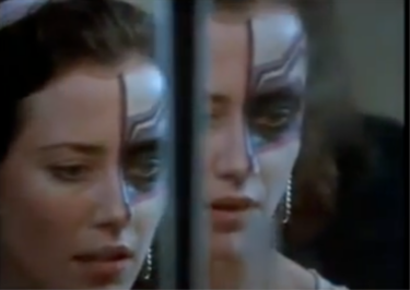 Two side-by-side photographs of the face of an actress in a television drama. She is a white woman with dark hair pulled or fixed up to reveal the whole face. The area around and above her left eye is painted with makeup. The makeup creates a large dark area around the eye, and the skin above is painted silvery grey with two wavy dark diagonal lines. 