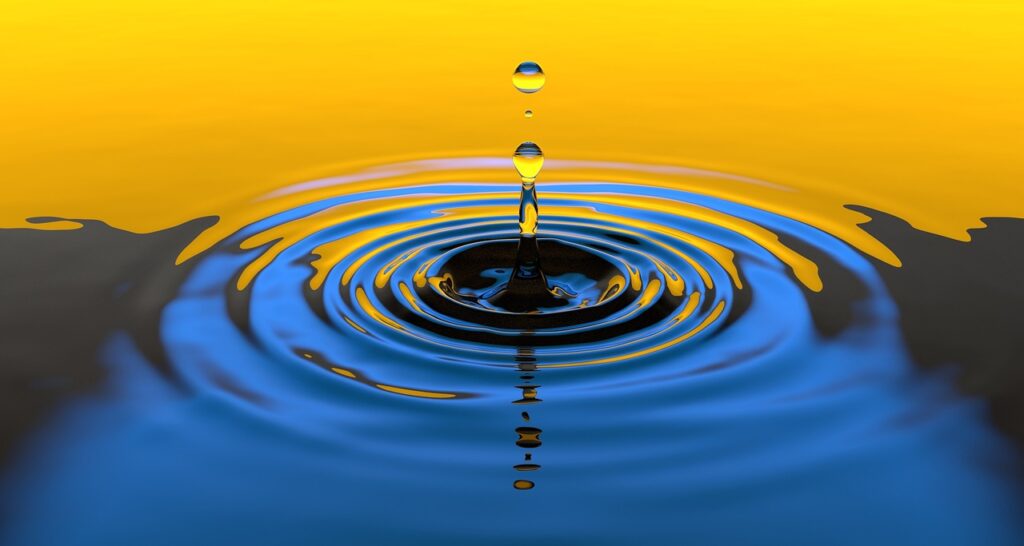A photograph of water droplets landing on calm water, producing radiating ripples on the water surface.