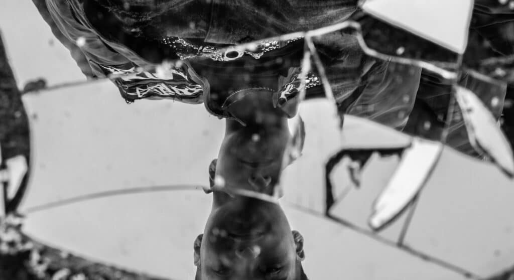 A black and white image of a person looking down at themselves into a broken mirror. The image is inverted, with the face at the bottom. They're wearing denim, logos and jewellery indicative of a counter-culture and have short hair.